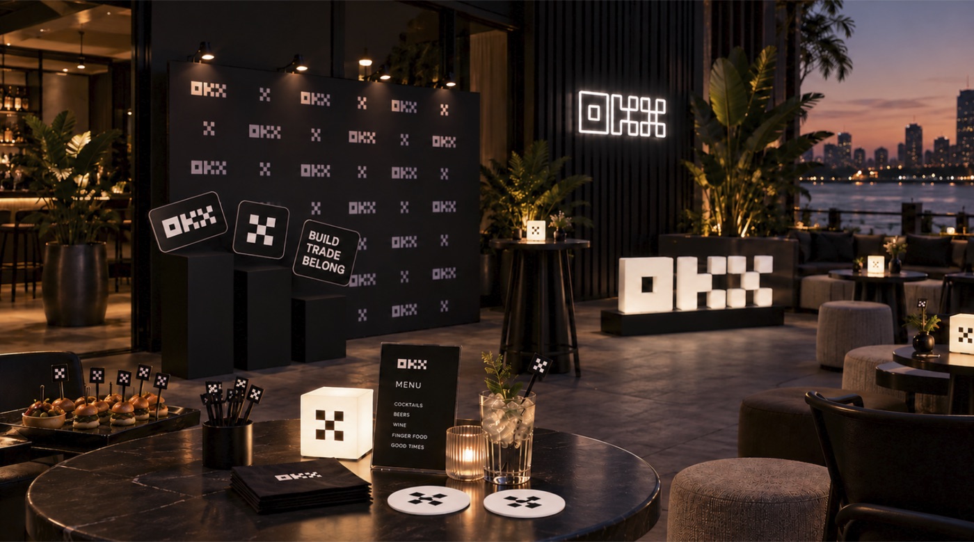 OKX branded event setup: step-and-repeat wall, illuminated OKX letters, branded menu, coasters and napkins on a terrace at dusk